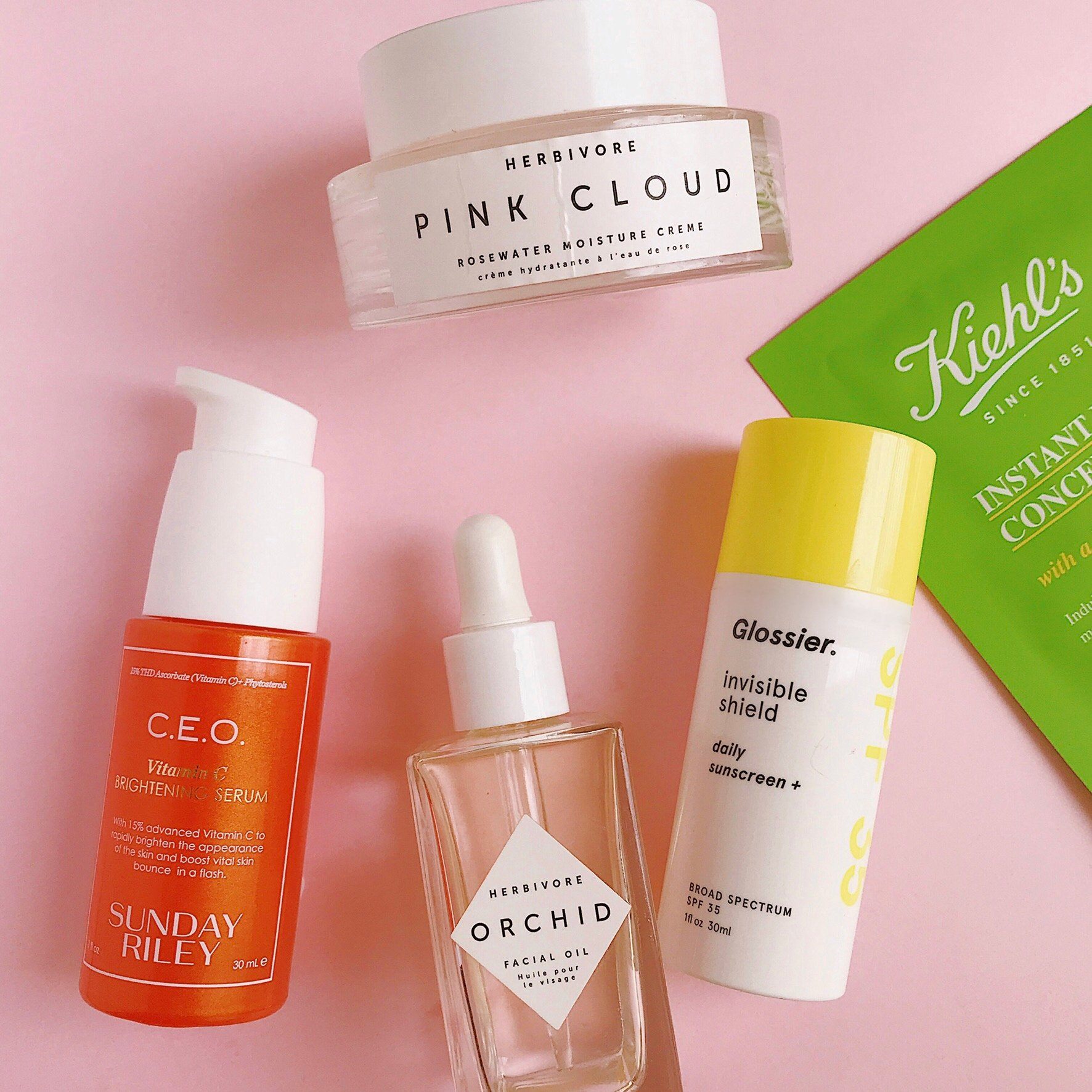 Skincare products against a pink background: C.E.O 15% Vitamin C Brightening Serum, Orchid Oil by Herbivore Botanicals, Pink Cloud Face Cream, glossier sunscreen and Kiehl's face mask.