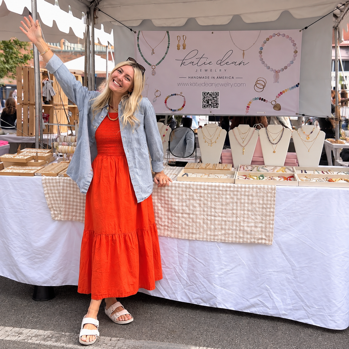 Katie Dean in an orange dress waving in front of a Katie Dean Jewelry booth displaying handmade necklaces, bracelets, and other jewelry at an outdoor market.