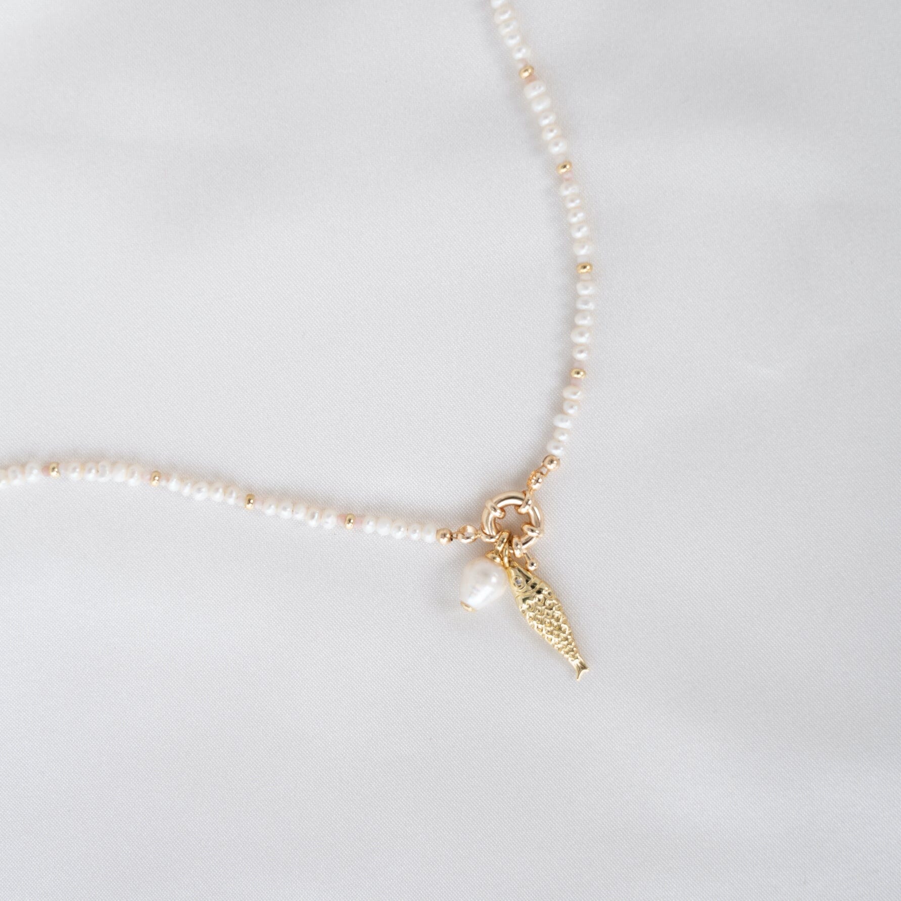 Pearl necklace with a small gold fish charm on a light gray background