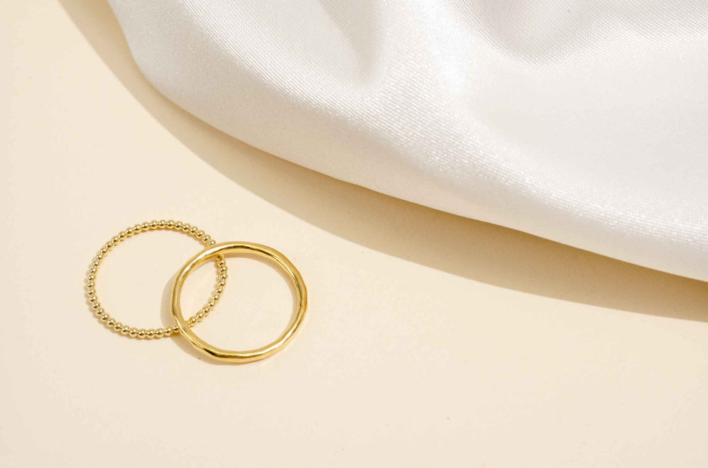 Two dainty gold stacking rings by Katie Dean Jewelry layered on top of each other against a light cream background, with a white satin cloth draped in the upper right corner of the image.