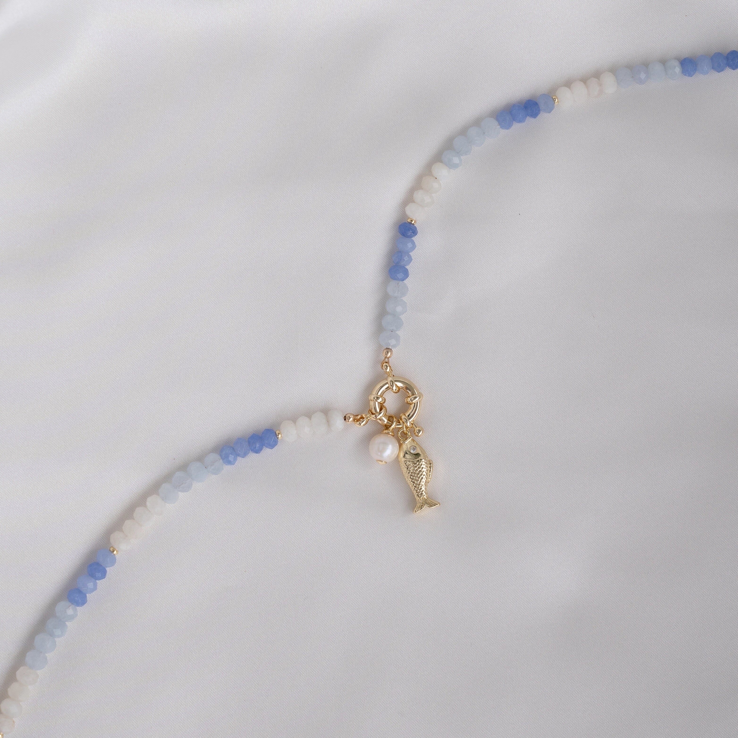Necklace with blue and white beads and a gold fish charm and freshwater pearl charm on a light gray background.