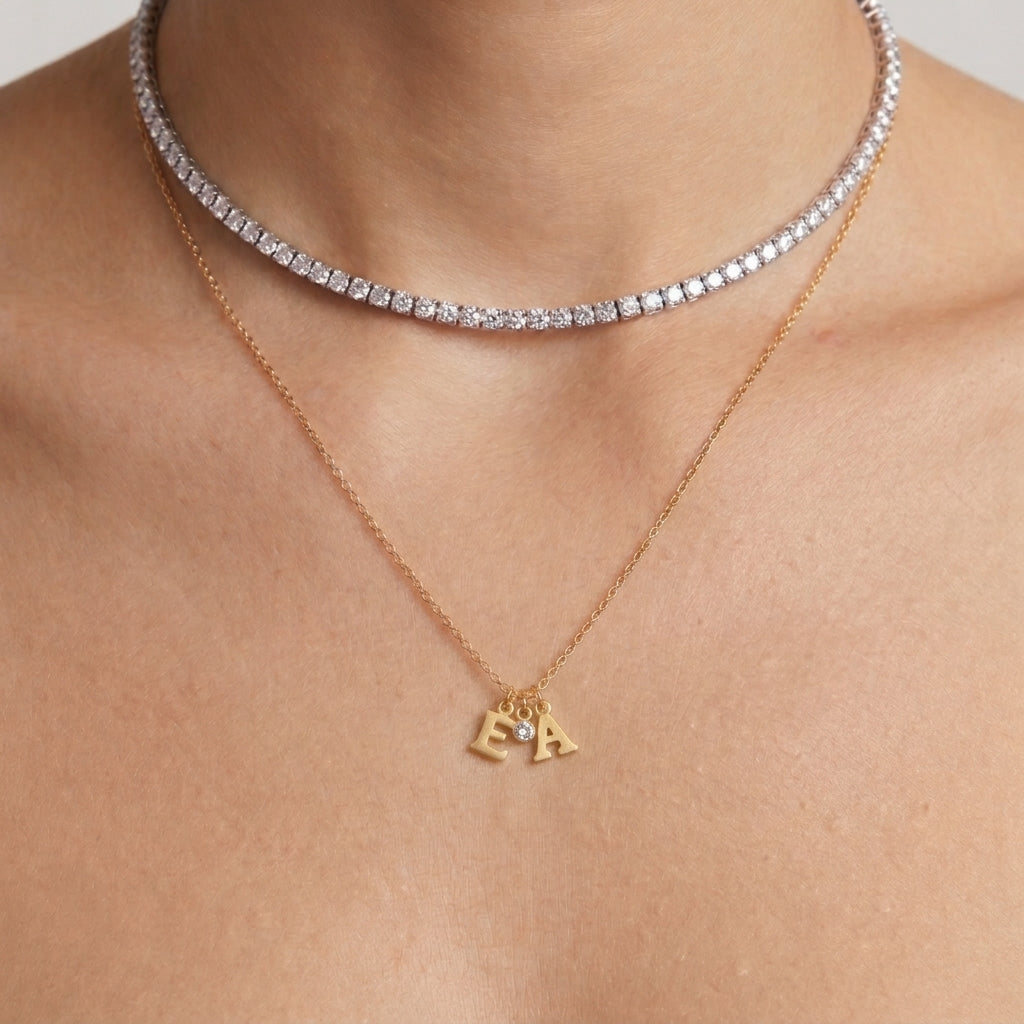 A close-up product photograph of a model wearing layered necklaces. The top layer is a sparkling diamond tennis choker, and the bottom layer is a delicate gold chain holding small, minimalist gold initial charms for the letters 'E' and 'A' flanking a tiny round-cut clear crystal birthstone charm. The jewelry is shown against a soft, professionally lit background.