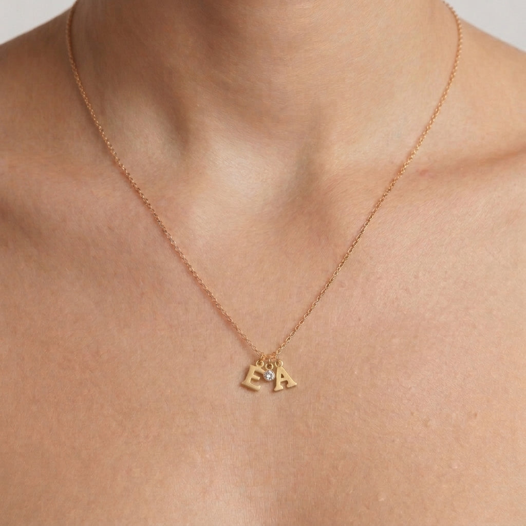 A close-up of a person's neck and upper chest wearing a delicate gold chain necklace with two small gold initial letters, 'E' and 'A', separated by a tiny clear stone.