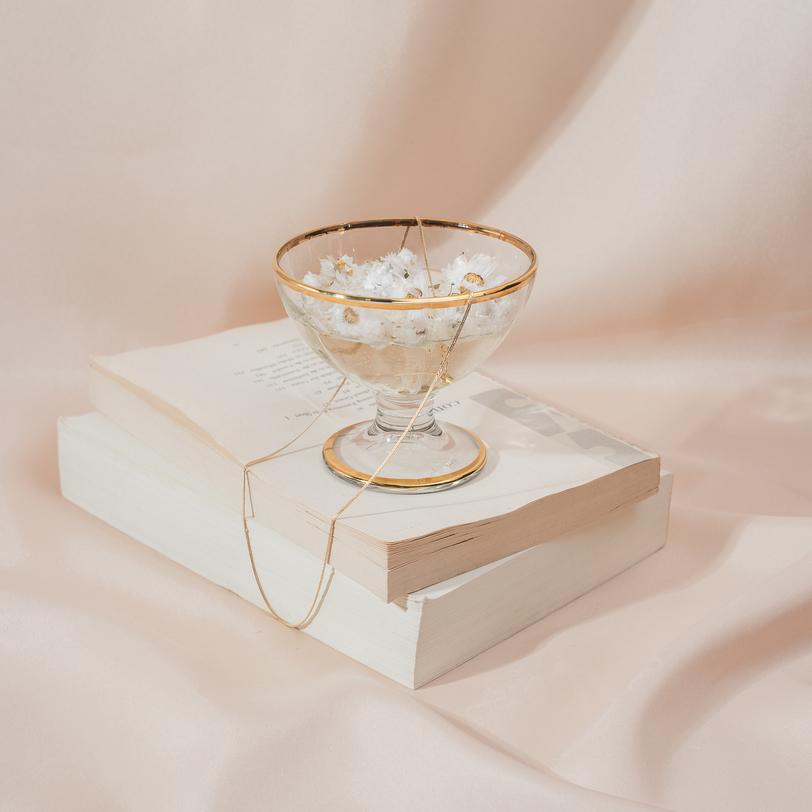 14k gold filled Herringbone Chain Necklace shown draped over a gold rimmed glass sitting on top of two books with a pink satin background, by Katie Dean Jewelry.