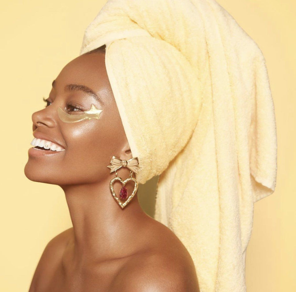 Picture of model in front of a yellow wall wearing a yellow towel around her head and gold eye masks by KNC beauty