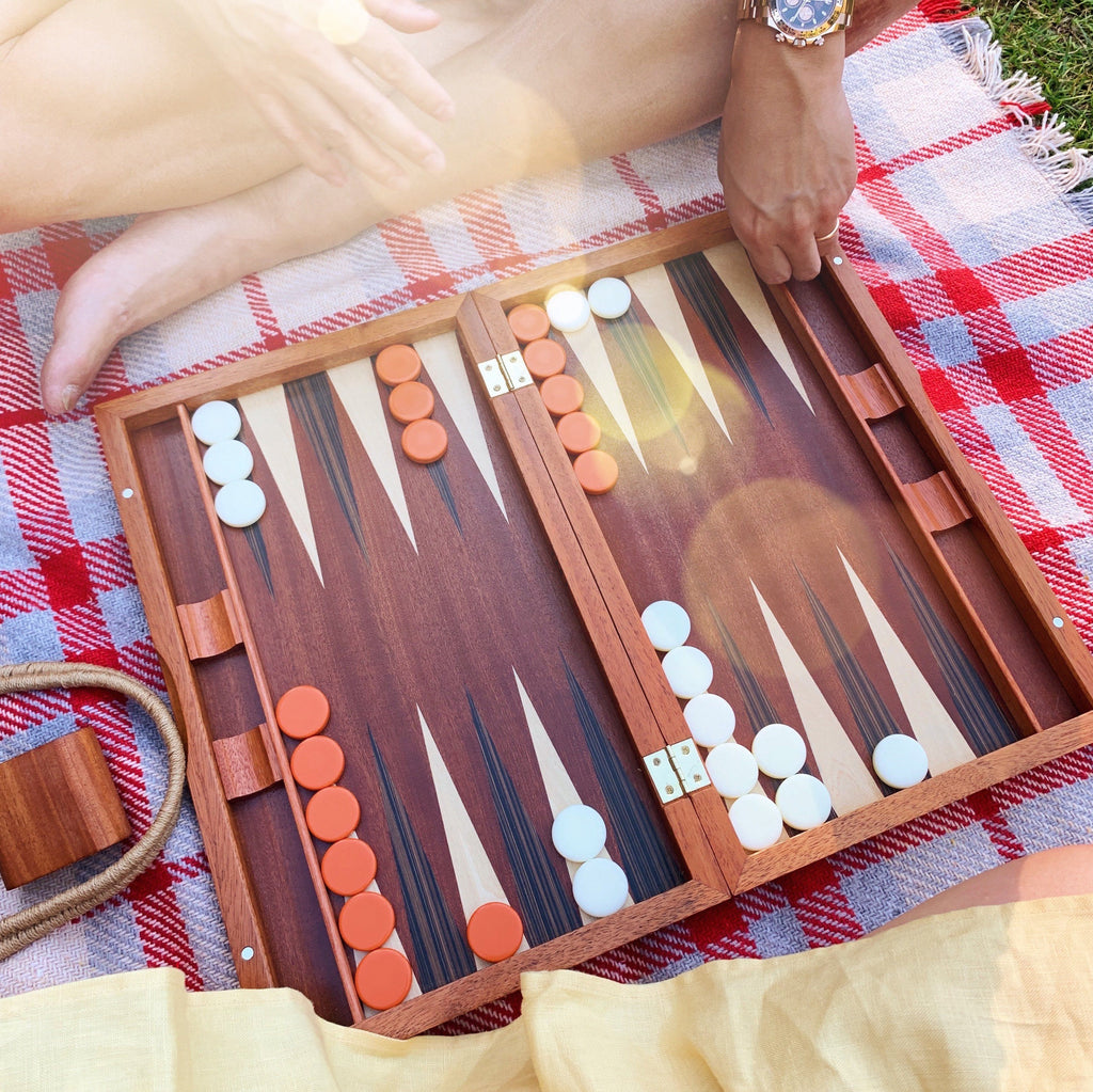 A wooden backgammon board sits atop a checkered picnic blanket. A yellow dress is visible on the bottom of the image and a hand coming into the frame is also visible as well as a ray of sunshine.