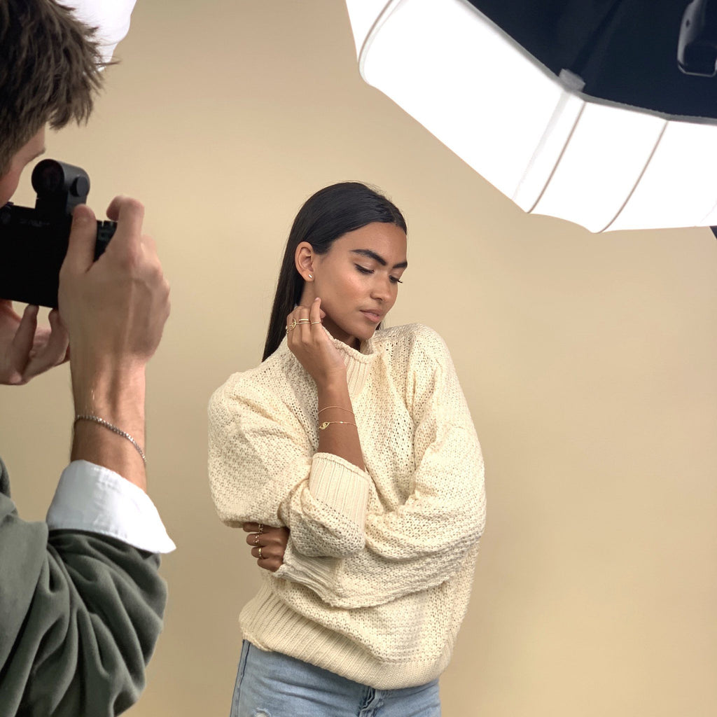 Image of a model posing with a cream turtleneck sweater and gold dainty stacking rings, bracelets and earrings on with a photographer taking a photo,Katie Dean Jewelry behind the scenes.