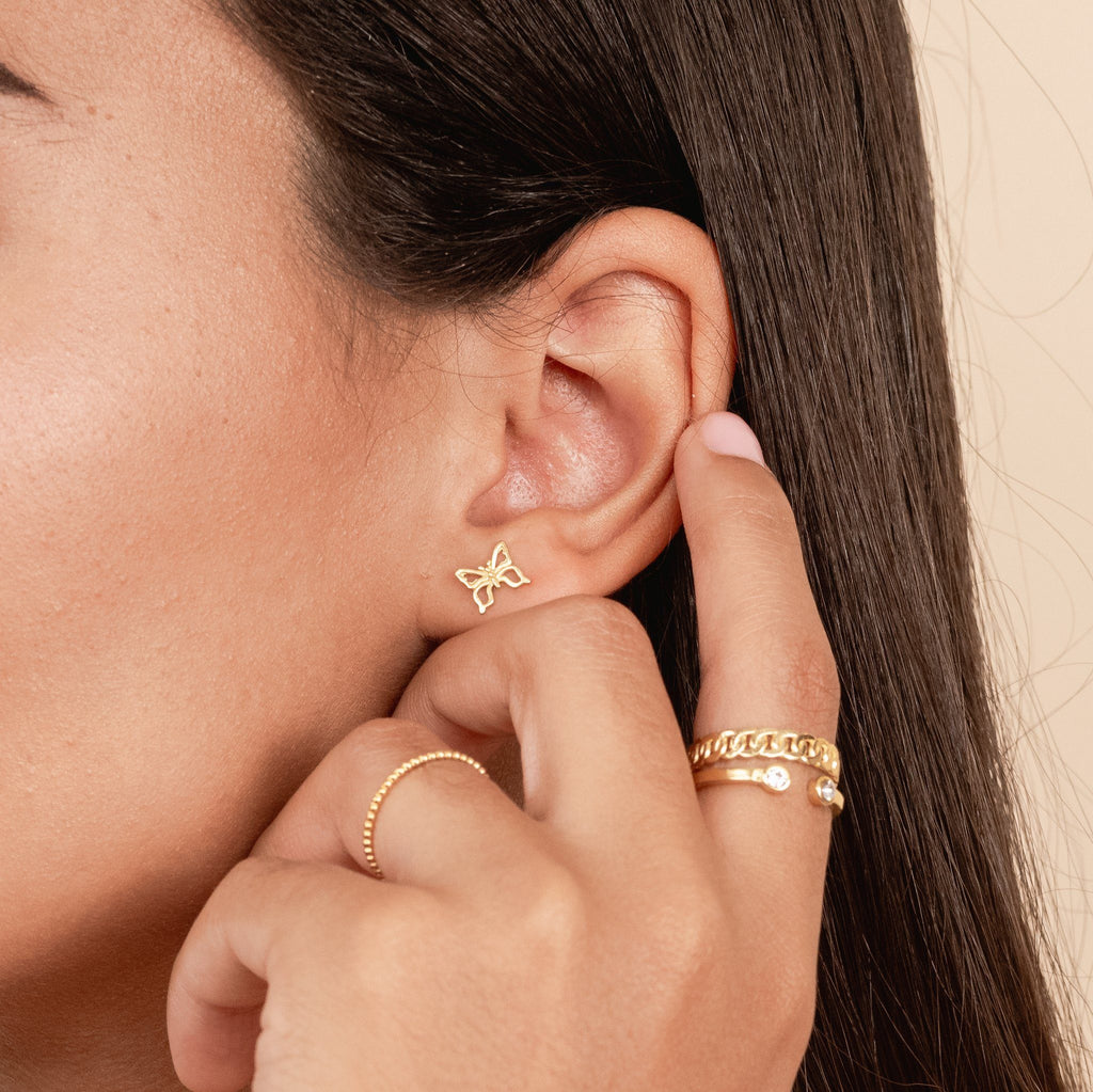 Dainty gold Butterfly Studs by Katie Dean Jewelry as seen on a model with her finger touching her ear wearing dainty gold stacking rings, handmade in the USA