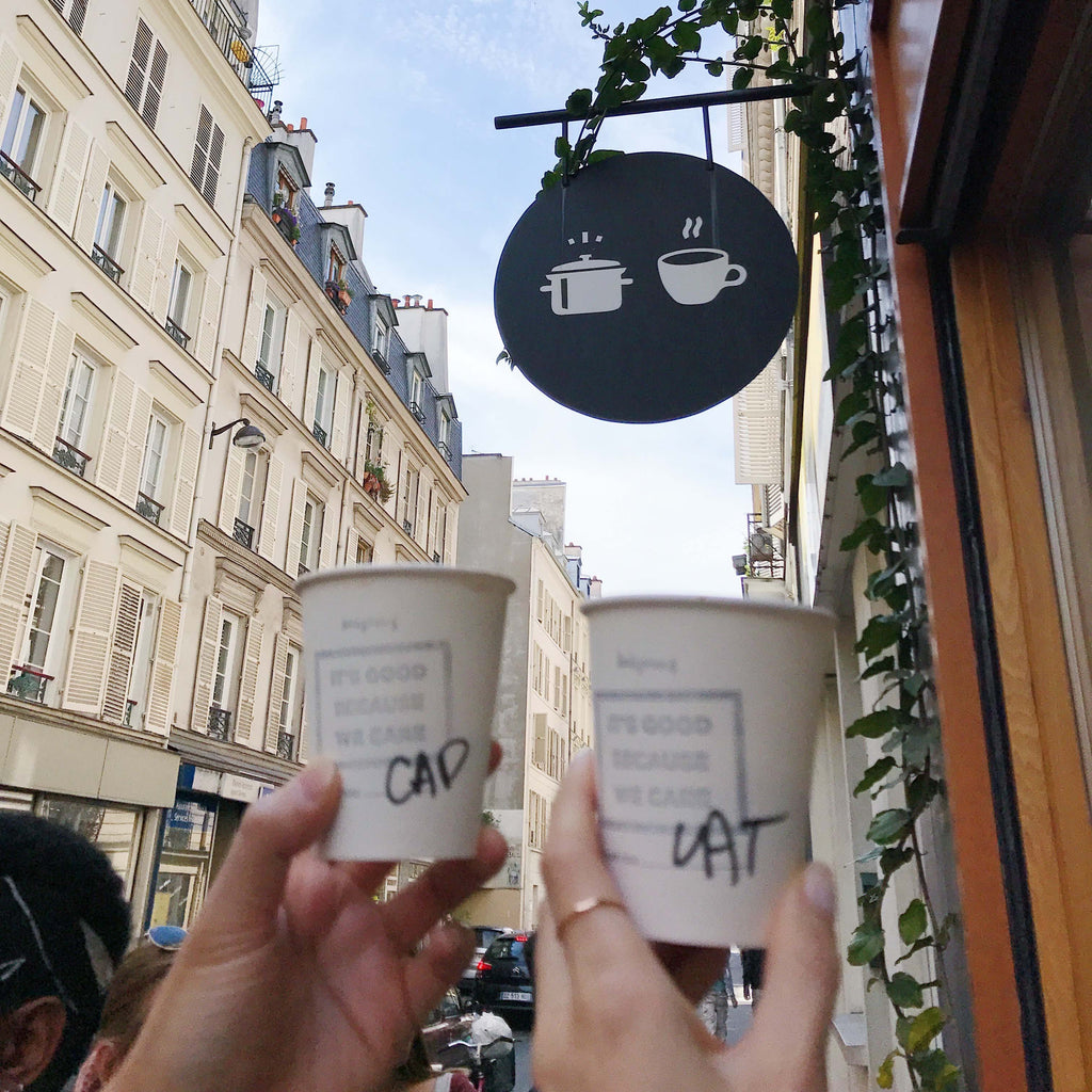 Cheerful toast with coffee cups under a quirky café sign in Paris, embodying the joy of shared moments and good beverages while showcasing a dainty gold band ring by Katie Dean Jewelry.