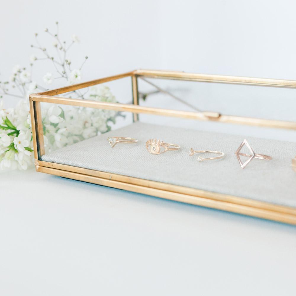 A gold-rimmed, glass display case with Katie Dean Jewelry rings displayed inside with white flowers next to the display.