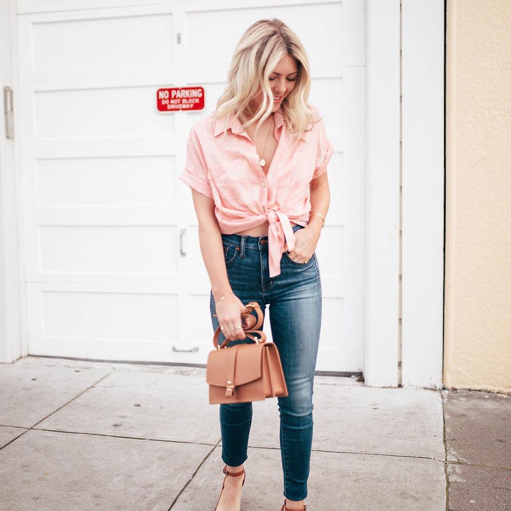 Katie standing outside holding a cute tan purse in a light pink crop top.
