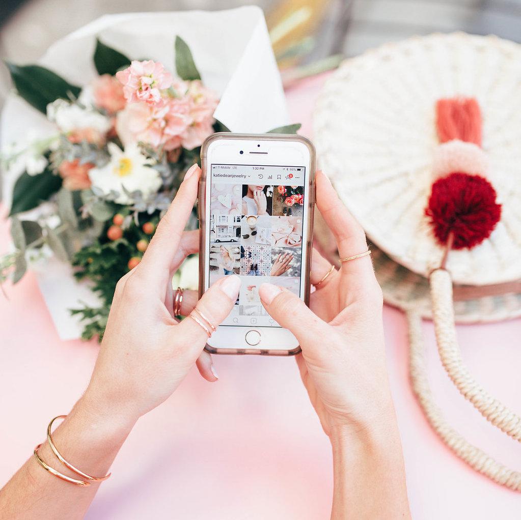 Two hands holding iphone showing Katie Dean Jewelry Instagram with flowers and straw bag on pink table. Gold rings and bangles on hands and wrists.