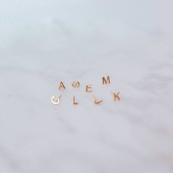 Gold initial stud earrings by Katie Dean Jewelry arranged on a marble surface, featuring letters A, S, E, M, L, and K.