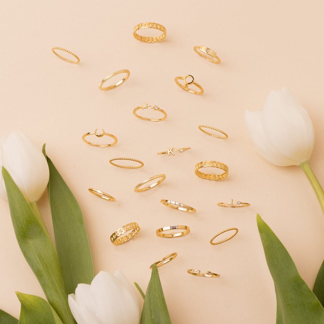 Assorted 14K gold-plated and gold-filled rings in various designs, including textured, smooth, and gem-set, elegantly displayed on a soft peach background with white tulips and lush green leaves.