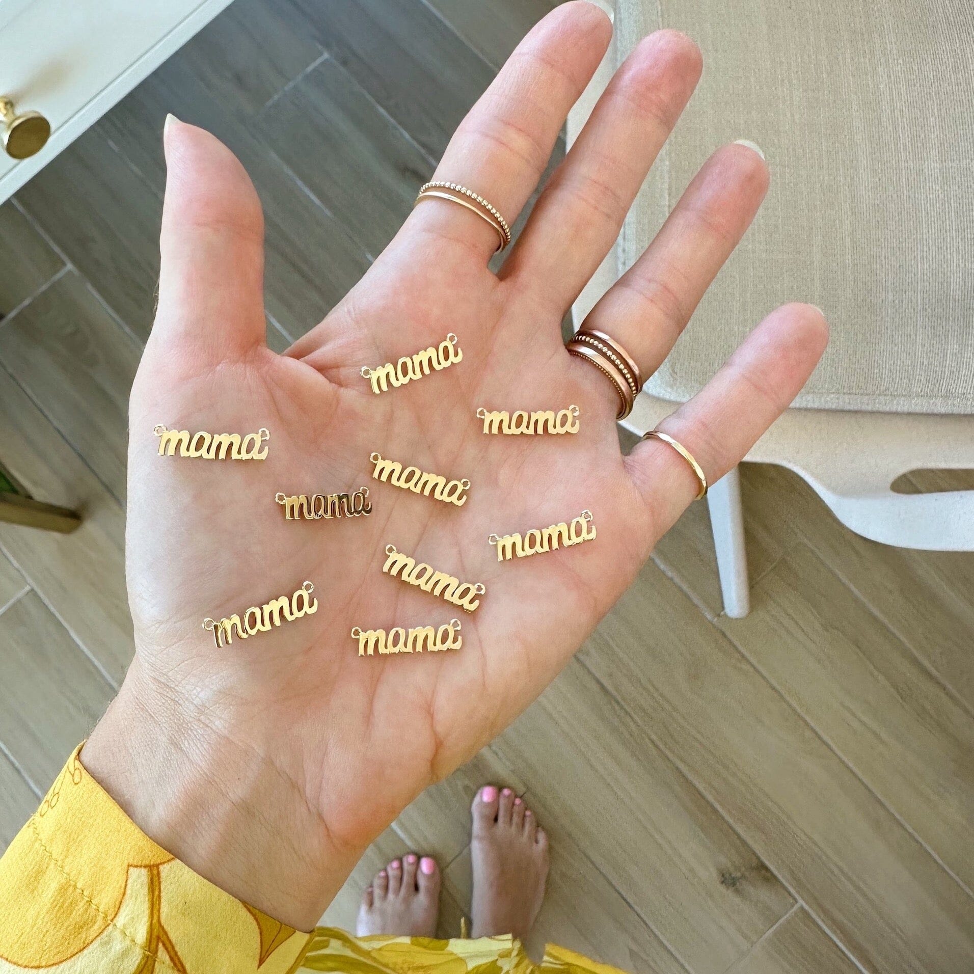 Gold Mama Charms on the palm of Katie's hand glowing from the natural light shining in. These dainty charms are made in America.