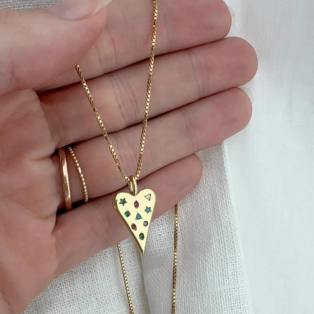 Gold heart-shaped necklace with colorful stones held between fingers on a light fabric background