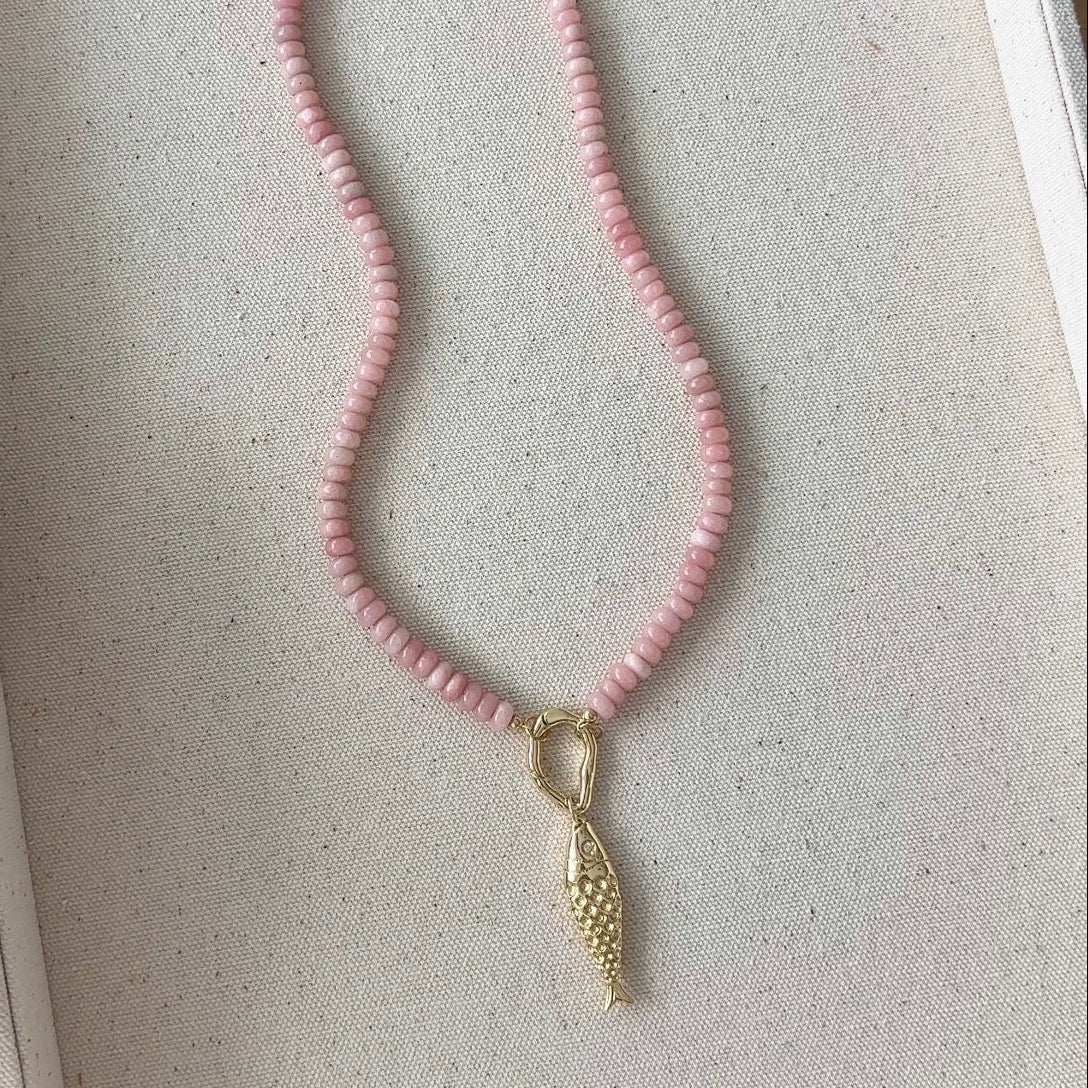 A pink beaded necklace with a gold fish pendant, displayed inside a linen jewelry box.
