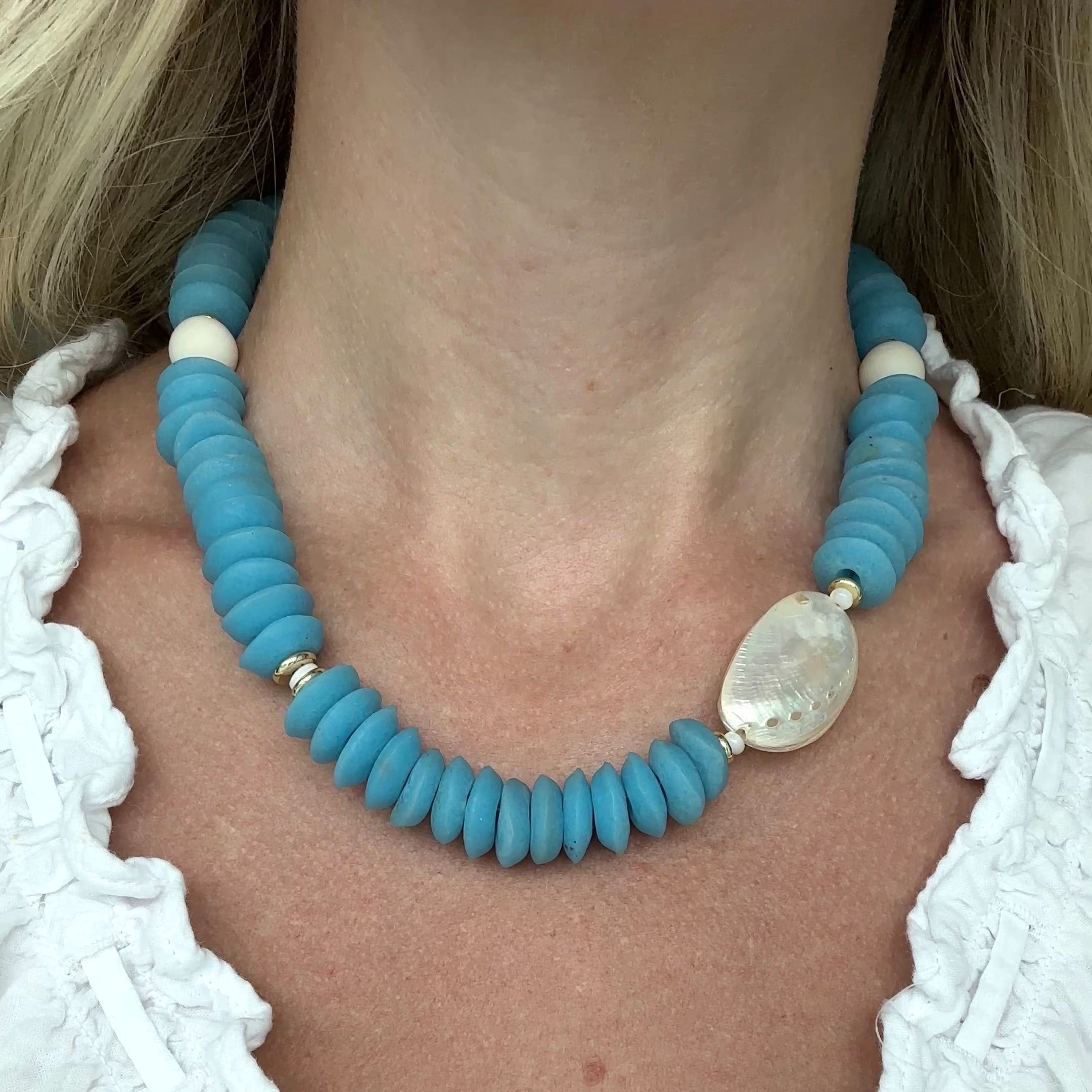 Necklace with blue beads and an abalone shell on a person wearing a white top.