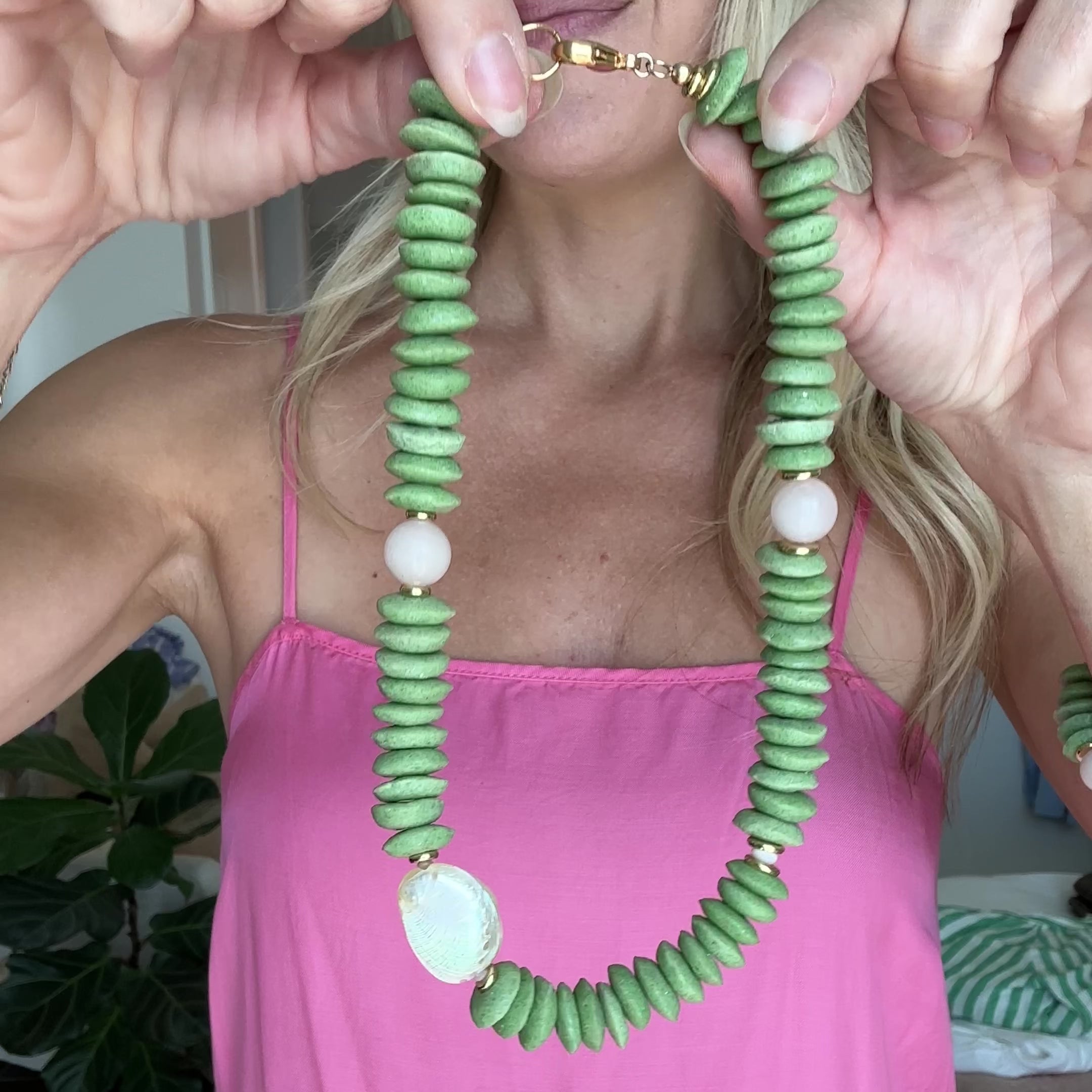 Necklace with green beads and white abalone shell and light pink quartz beads with gold elements being held up by a persons hands.