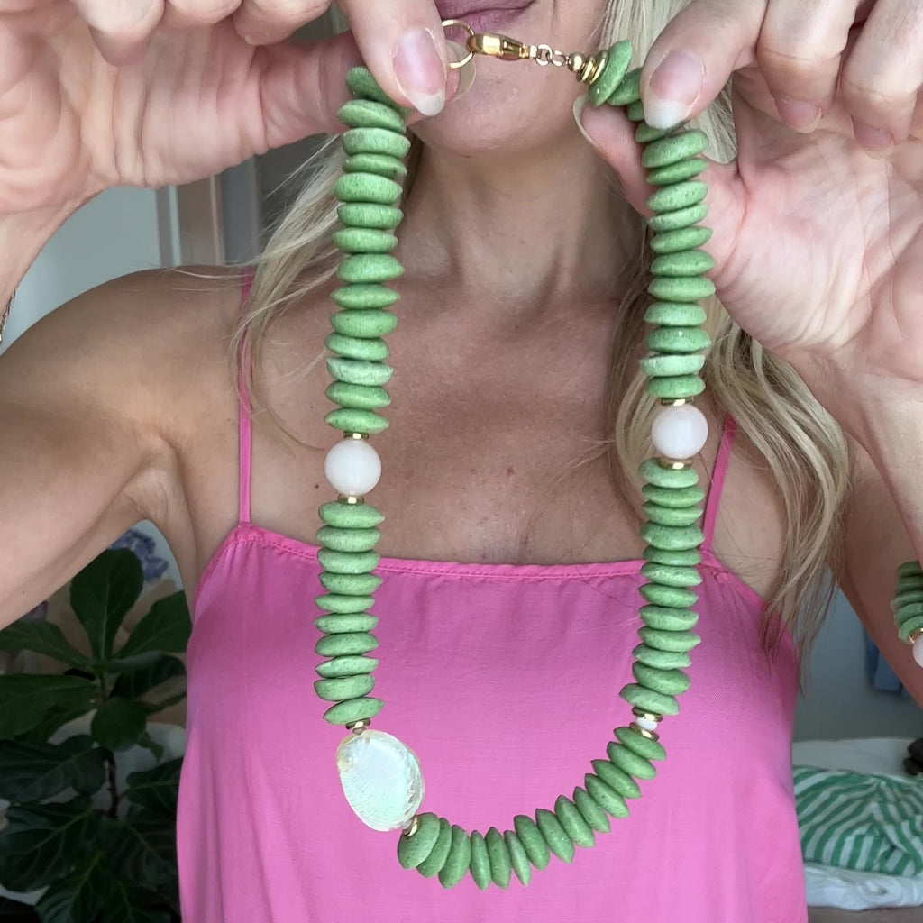 Necklace with green beads and white abalone shell and light pink quartz beads with gold elements being held up by a persons hands.