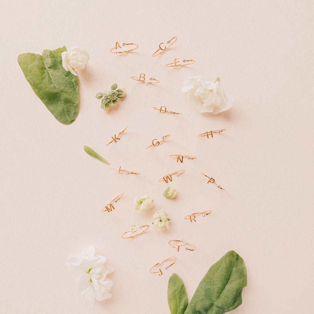 Gold dainty Initial Rings shown on a pink background with white flowers and green leaves