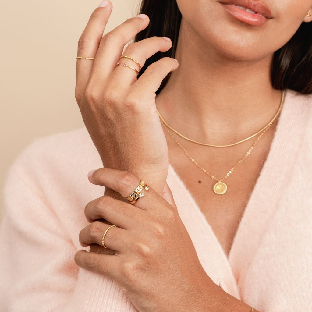 Model wearing pink sweater and holding up her hands, wearing dainty gold stacking rings made by Katie Dean Jewelry. Showing the Hammered Band Ring, Birthstone Ring, Figaro Chain ring, Two Gem Ring and Beaded Ring.