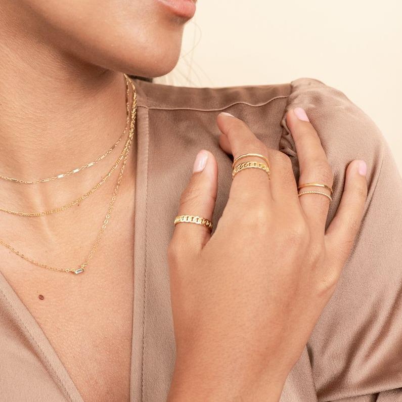 Model wearing brown silk top, looking to the side, wearing the gold Linked Choker, Figaro Chain Choker and Baguette Necklace. On her fingers she is wearing the gold Figaro Chain Rings and Hammered Band and Beaded stacking Rings. Handmade in California by Katie Dean Jewelry.