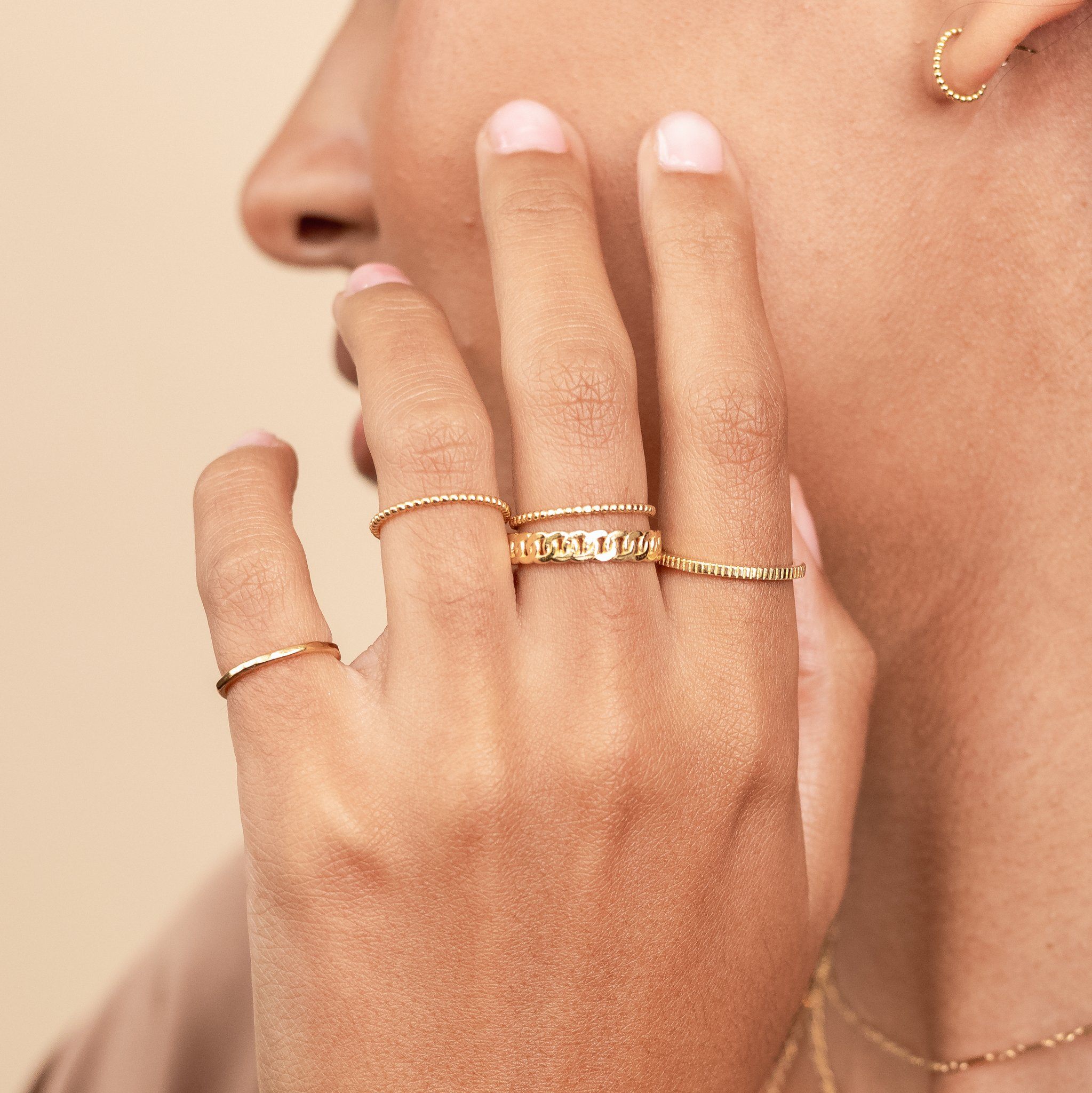 Model holding up her hand to her face, wearing dainty gold stacking rings made by Katie Dean Jewelry. Showing the Hammered Band Ring, Coin Ring, Figaro Chain ring, and Beaded Ring.