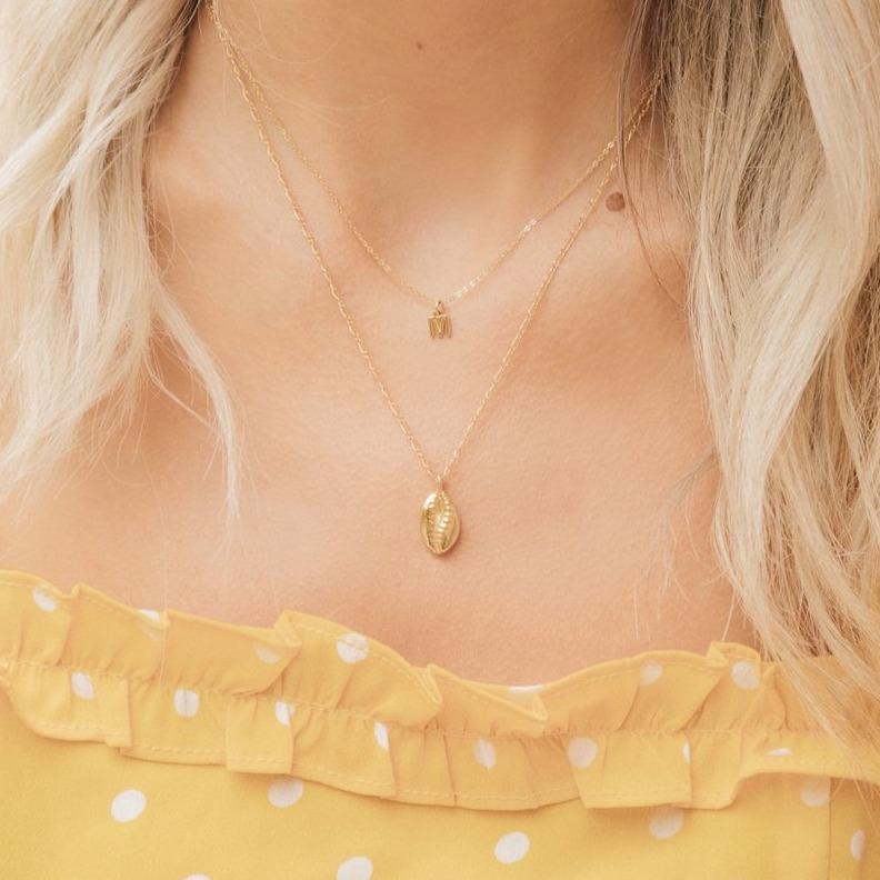 Puka Shell Necklace, gold and dainty layered with an Initial "M" Necklace also gold. Model has blonde hair and is wearing a yellow dress with white polka dots.