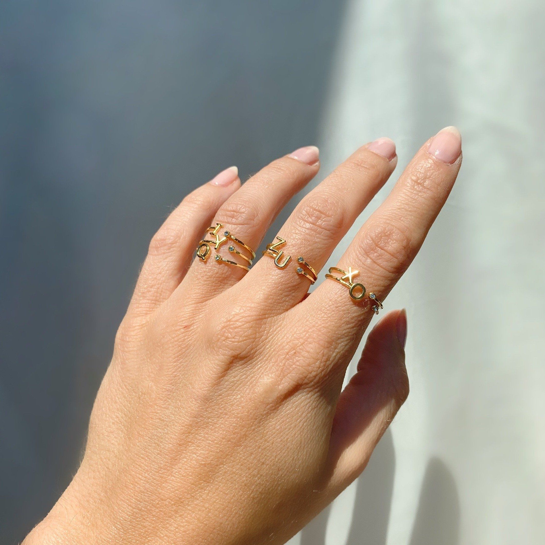 Dainty gold Initial Rings i, o, q, u, x, y, z sparkling in the sunlight as shown on a hand with a piece of white satin behind.