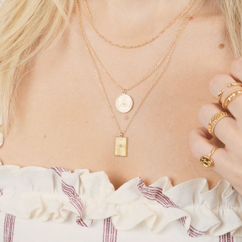Model wearing three layered gold dainty necklaces. Top is a plain chain, middle is a circular charm necklace, bottom is a rectangle charm necklace. All gold in color.
