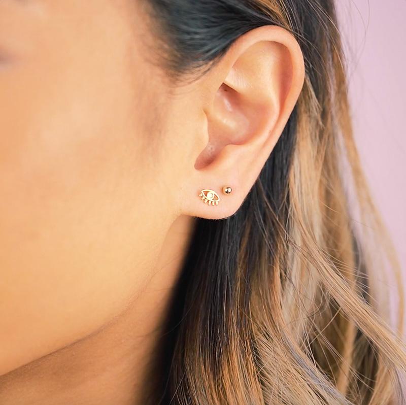 Image of the left side of the face showing an ear with two piercings, one with a gold eye earring and another with a small ball bead earring.